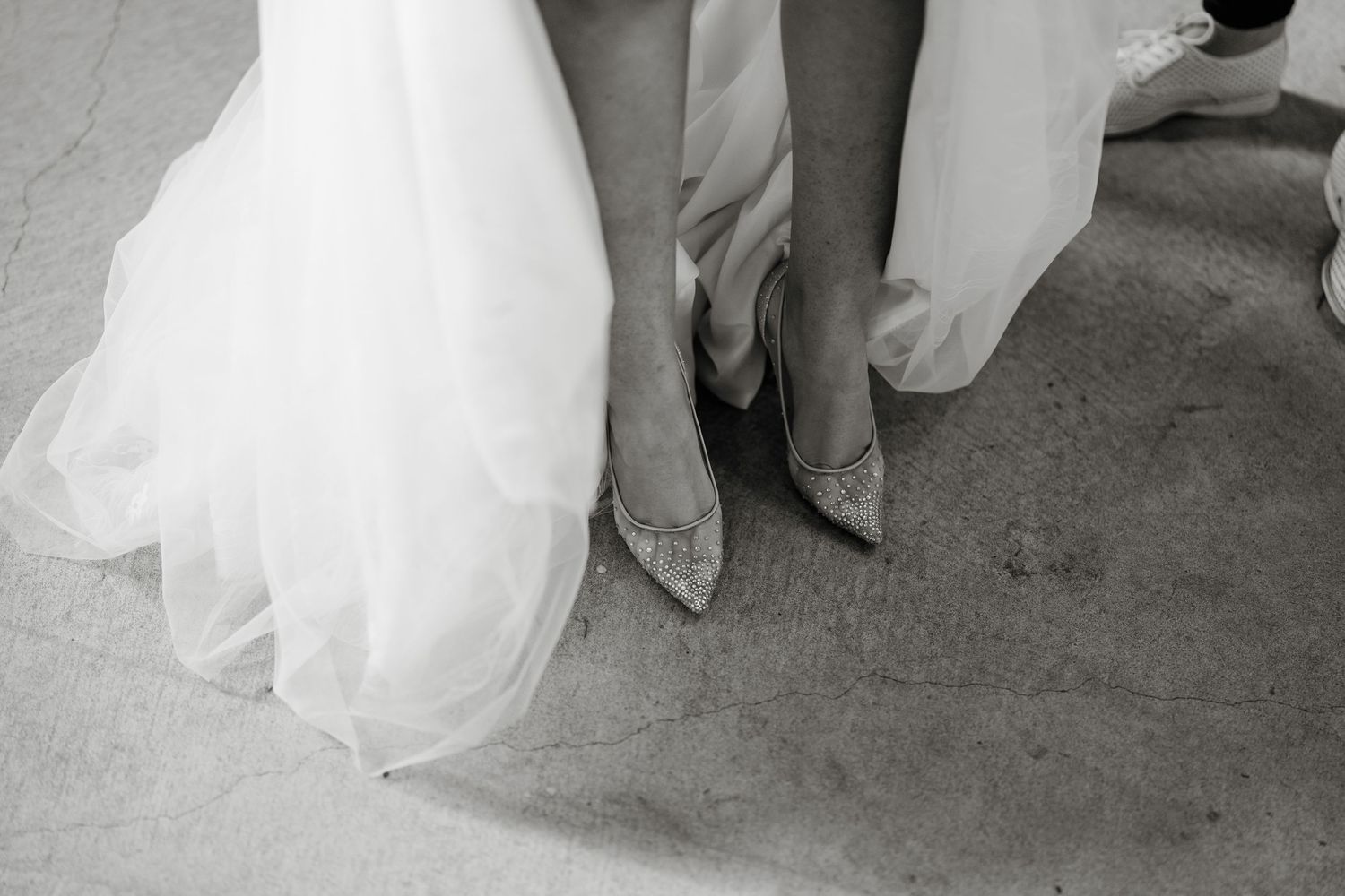 Black and white photo of a bride in a white gown wearing sparkly heels on a concrete floor.