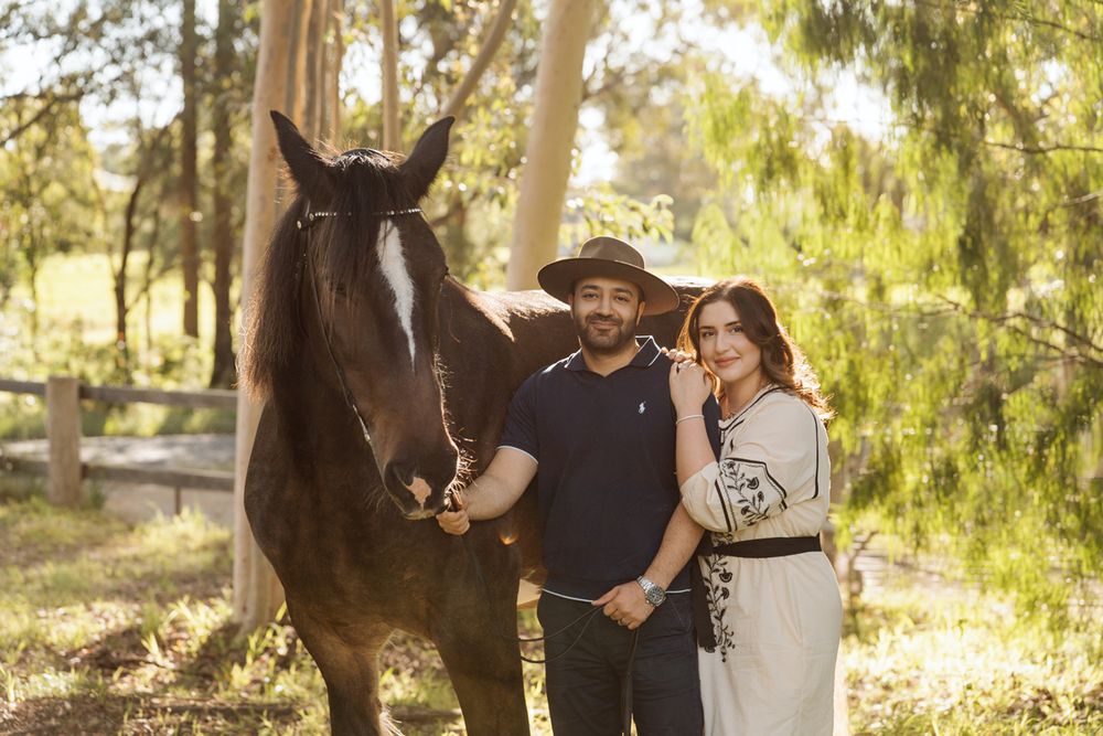 Couple in hats pose with dark horse in sunny tree-lined paddock, woman in embroidered dress, man in navy polo in sydney