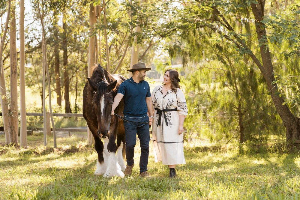 Couple walking with a Clydesdale horse through a sunlit green meadow surrounded by tall trees in sydney