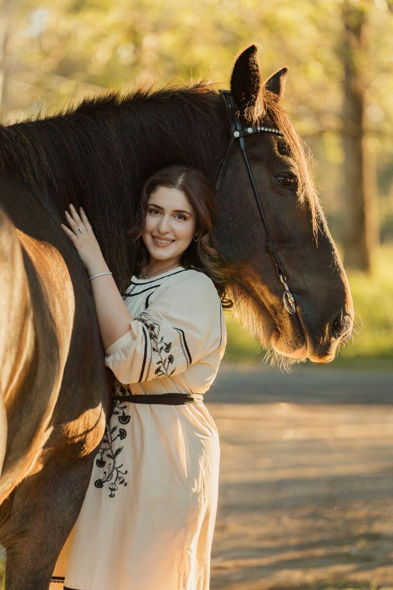 Young woman in embroidered cream dress smiling beside a dark brown horse in a sunlit forest setting in sydney