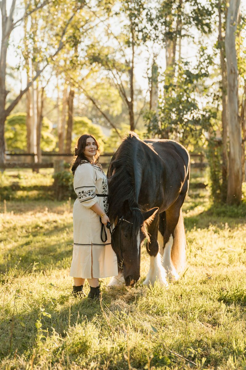 Woman in embroidered dress stands beside grazing dark horse in sunlit paddock surrounded by eucalyptus trees in sydney