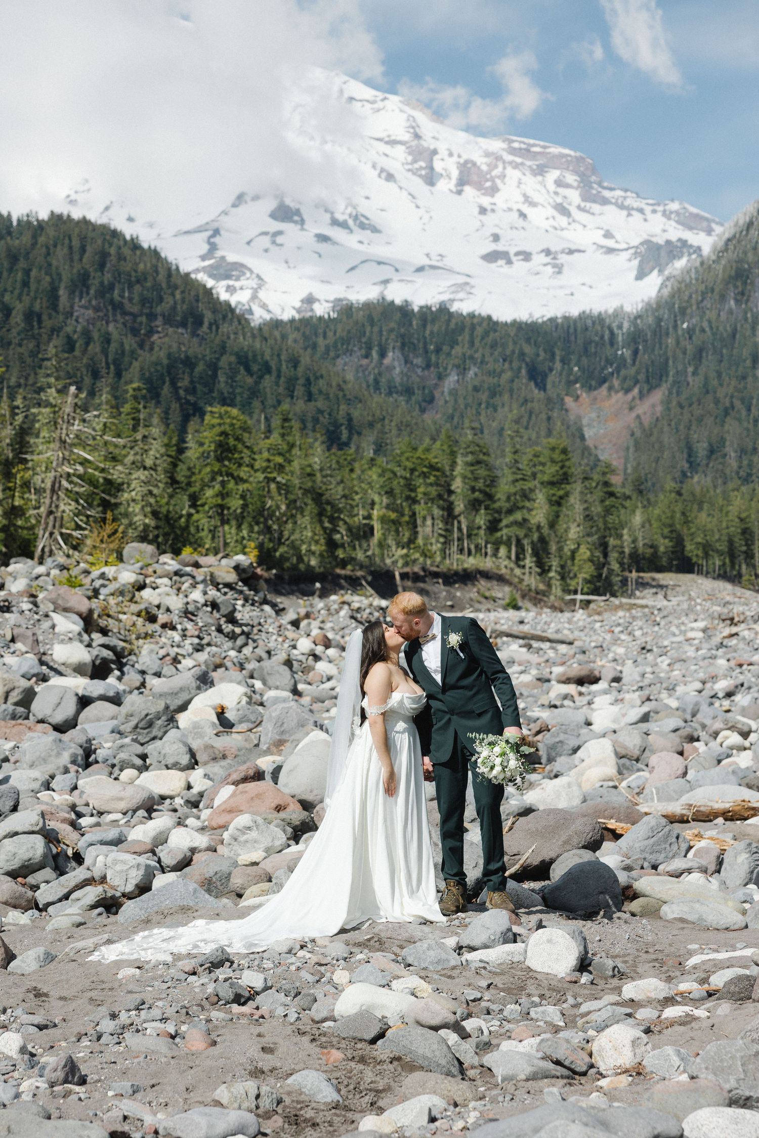 Bride and groom kissing on rocky terrain with snow-capped Mount Hood and evergreen forest backdrop.