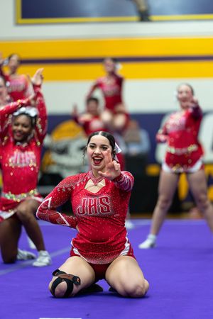A cheerleader in a red JHS uniform kneels on a purple mat, smiling and gesturing during a competition.