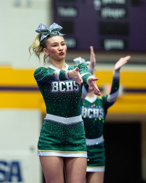 BCHS cheerleaders in green rhinestone uniforms perform at a competition, with a blonde flyer striking a pose in the foreground.