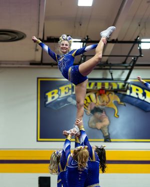 Blue and gold cheerleader executes a high leg hold stunt lifted by teammates at indoor competition.