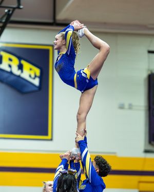 A cheerleader in a blue and gold uniform performs a high lib stunt, extending her leg overhead while being held aloft by teammates.