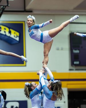 A cheerleader in a light blue uniform executes a high kick stunt while being elevated by teammates during a cheerleading competition.