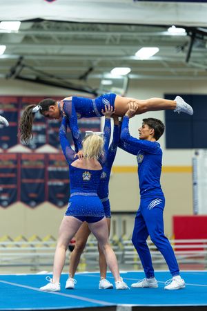 Cheerleaders in blue uniforms perform a stunt lifting a flyer horizontally at an indoor competition.