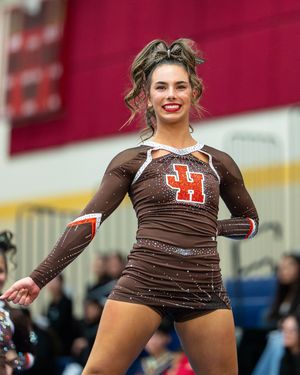 A smiling cheerleader in a brown bedazzled JH uniform performs during a competitive cheerleading competition.