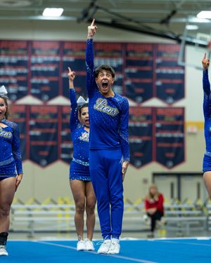 A male cheerleader in a royal blue Lions uniform energetically points upward while performing with his squad during a competition.