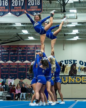Cheerleaders in blue uniforms performing a high stunt lift during an indoor competition near state champion banners.