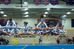 Cheerleading squad in light blue uniforms performs synchronized jumps at indoor cheer competition.