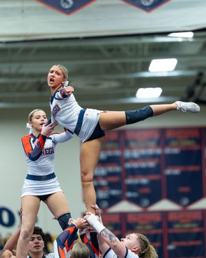 Female cheerleader in white and navy uniform executing a high kick stunt lifted by teammates at a competition.