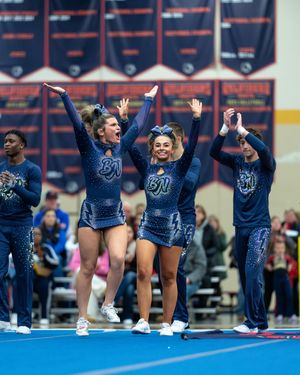 Three cheerleaders in navy BN uniforms cheer enthusiastically on a competition floor.