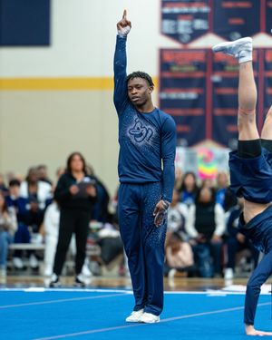 A male cheerleader in a navy uniform raises one finger skyward while teammates perform stunts behind him at a cheerleading event.
