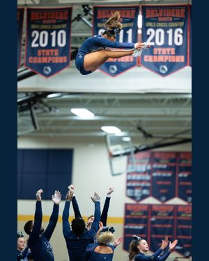 Cheerleader performs a high-flying tuck jump stunt while teammates reach up to catch her in a school gymnasium.