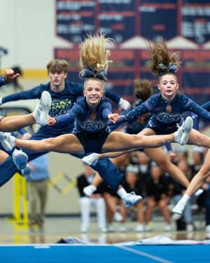 Cheerleaders in navy sparkle uniforms performing synchronized toe-touch jumps at an indoor cheerleading competition.