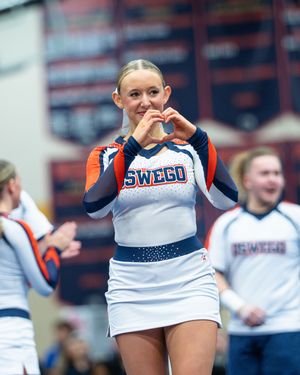 Smiling Oswego cheerleader in white and orange uniform makes heart shape with hands at competition.