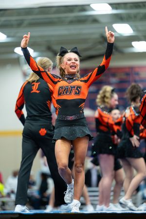 Cheerleader in orange CATS uniform performs energetically with arms raised at indoor competition.
