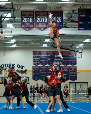 A cheerleader in a black and orange uniform is tossed high in the air during a competition stunt.