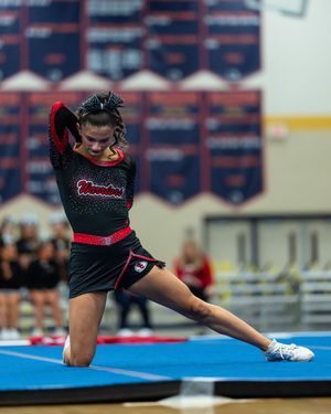 Cheerleader in black and red uniform performs a solo routine on blue mat at competition.