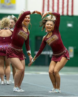 Two cheerleaders in maroon uniforms perform synchronized dance moves at an indoor cheerleading competition.