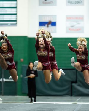 Three cheerleaders in maroon uniforms leap simultaneously with raised fists during a competitive cheer routine.