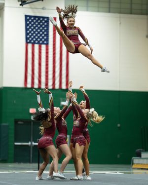 A cheerleader in maroon is tossed high into the air by teammates beneath an American flag indoors.