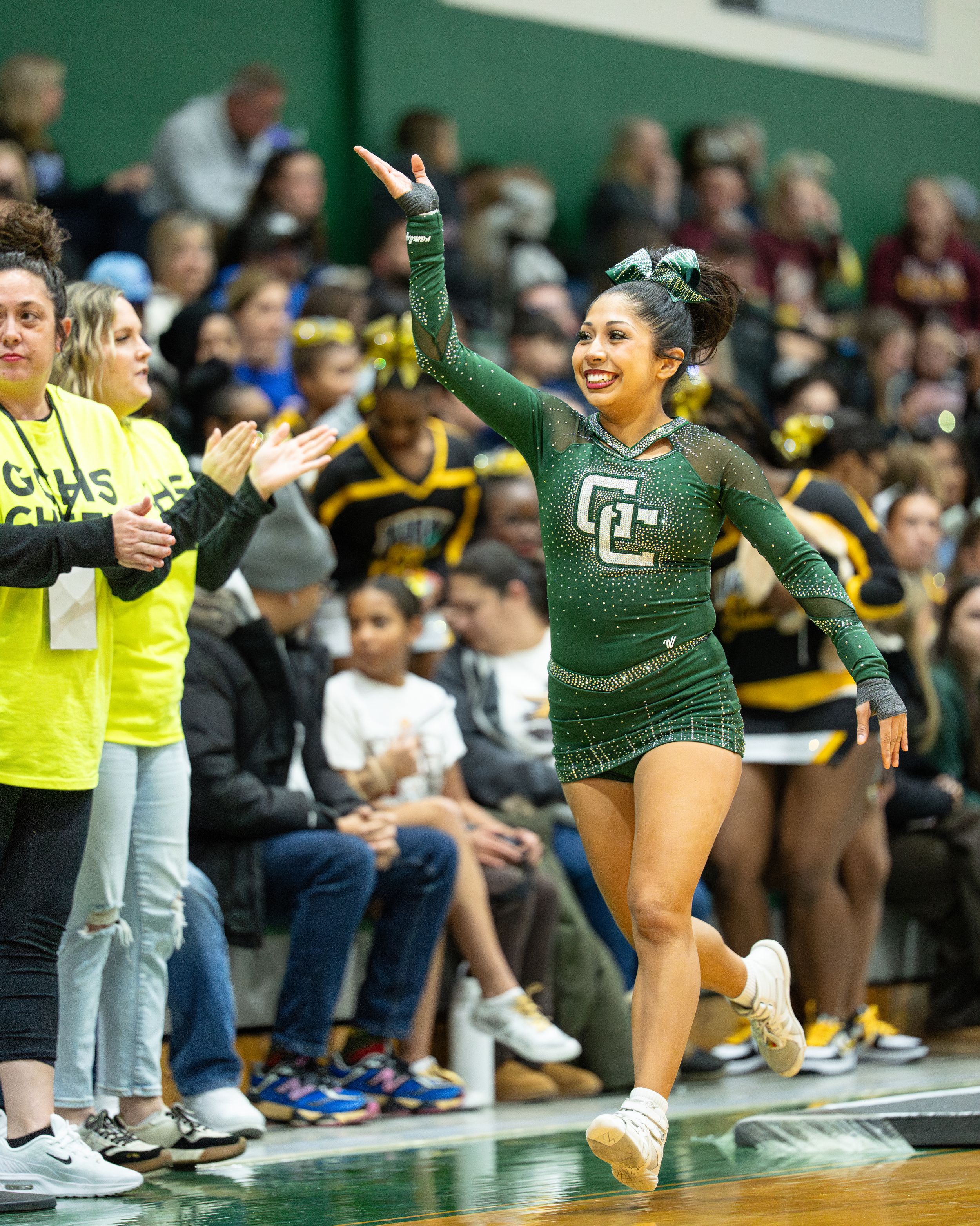 A cheerleader in a green uniform performs at a basketball game, raising her hand while teammates and crowd cheer.