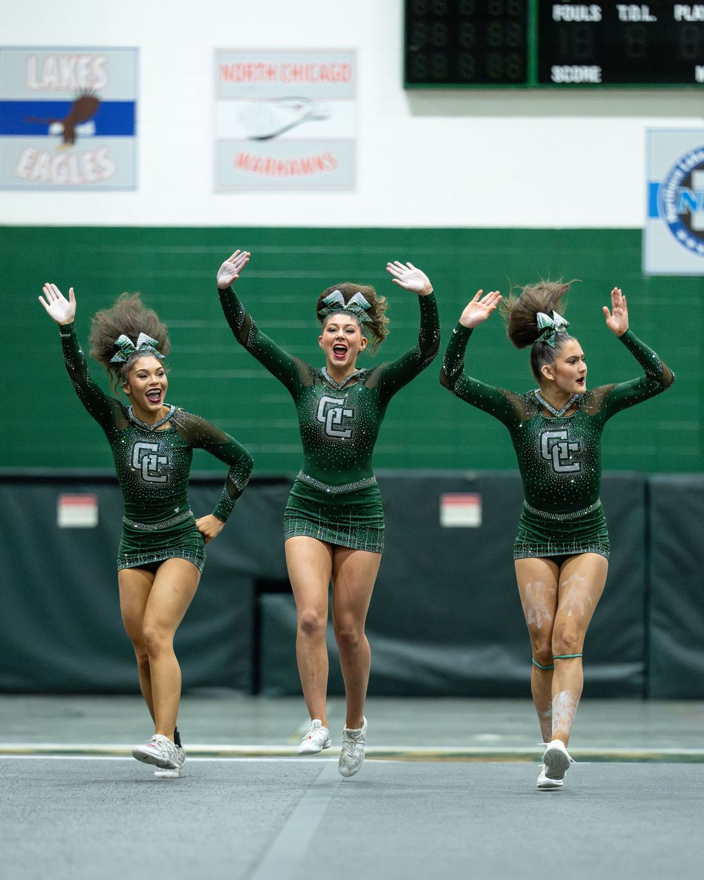 Three cheerleaders in green plaid uniforms jump and cheer energetically during a competition.