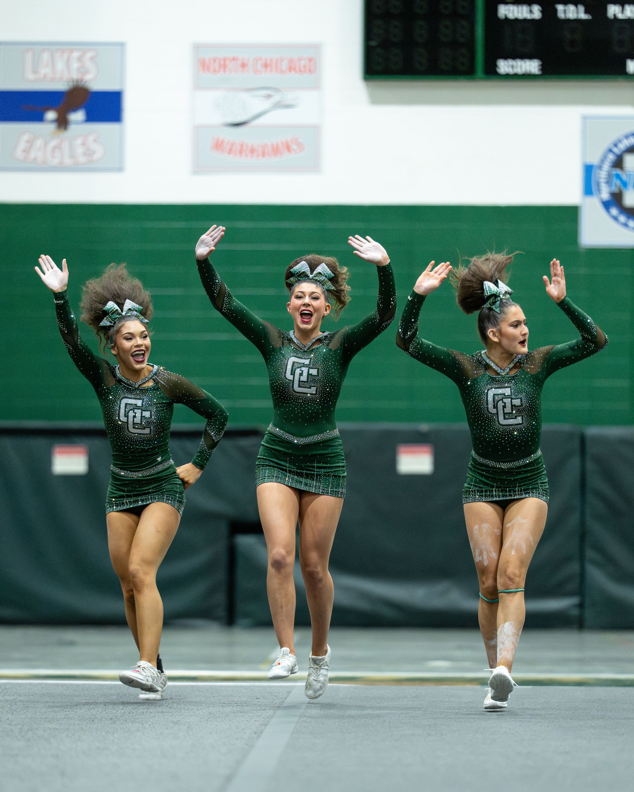 Three cheerleaders in green plaid uniforms jump and cheer energetically during a competition.