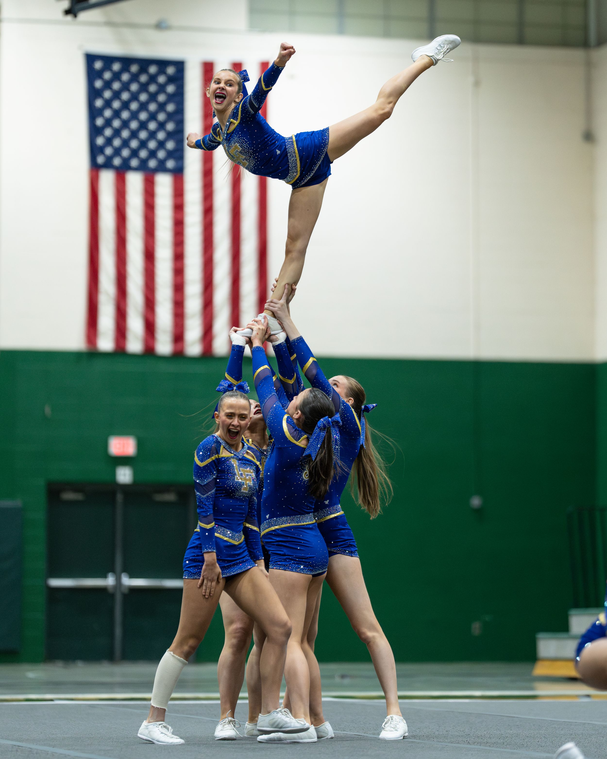 Cheerleaders in blue uniforms perform a stunt, lifting a flyer high in a gym with an American flag backdrop.