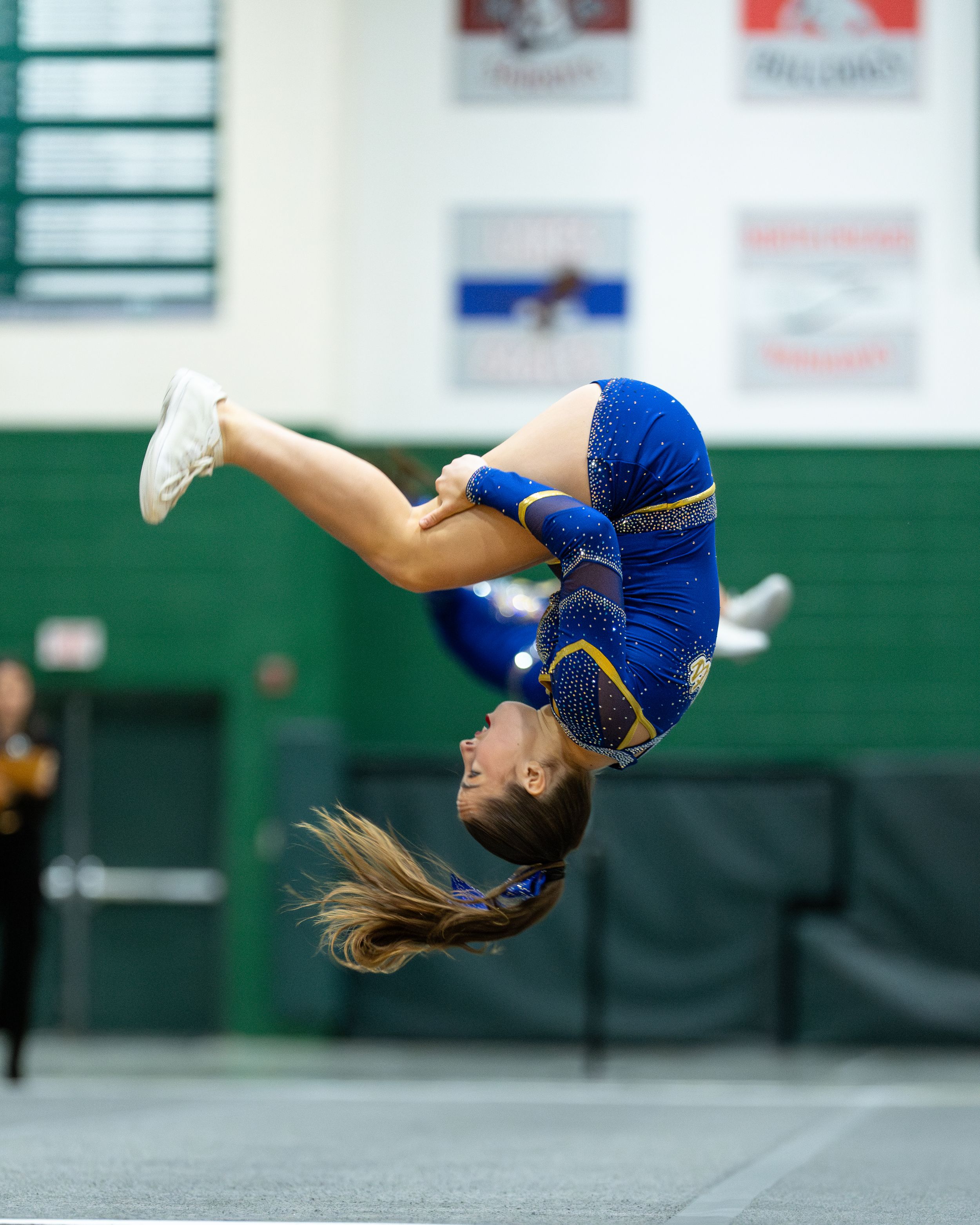 Cheerleader in blue sparkly uniform performs a backflip mid-air during a cheerleading competition in a gymnasium.