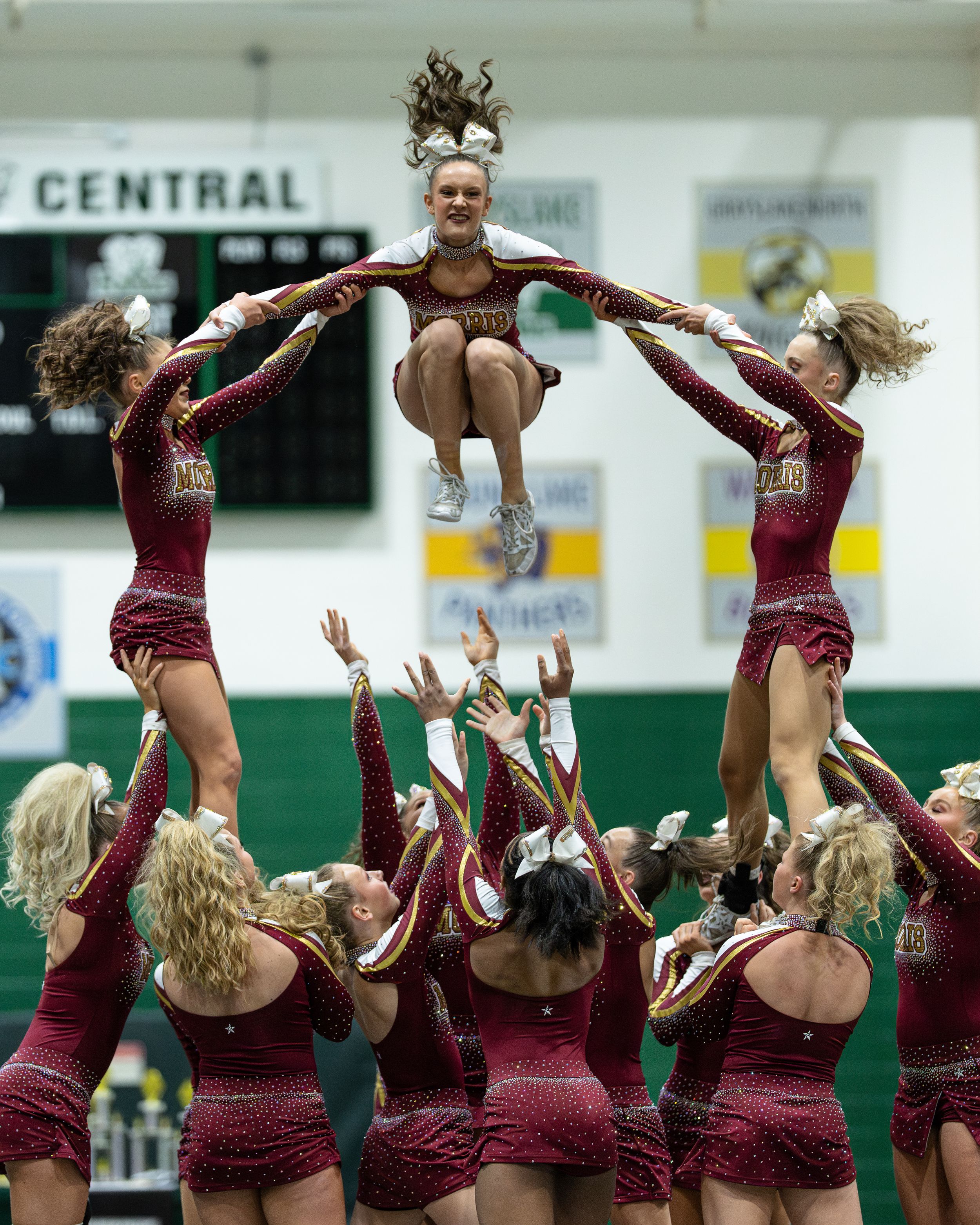 Cheerleaders in maroon uniforms perform a stunning pyramid stunt, lifting a flyer high in the air during a competition.