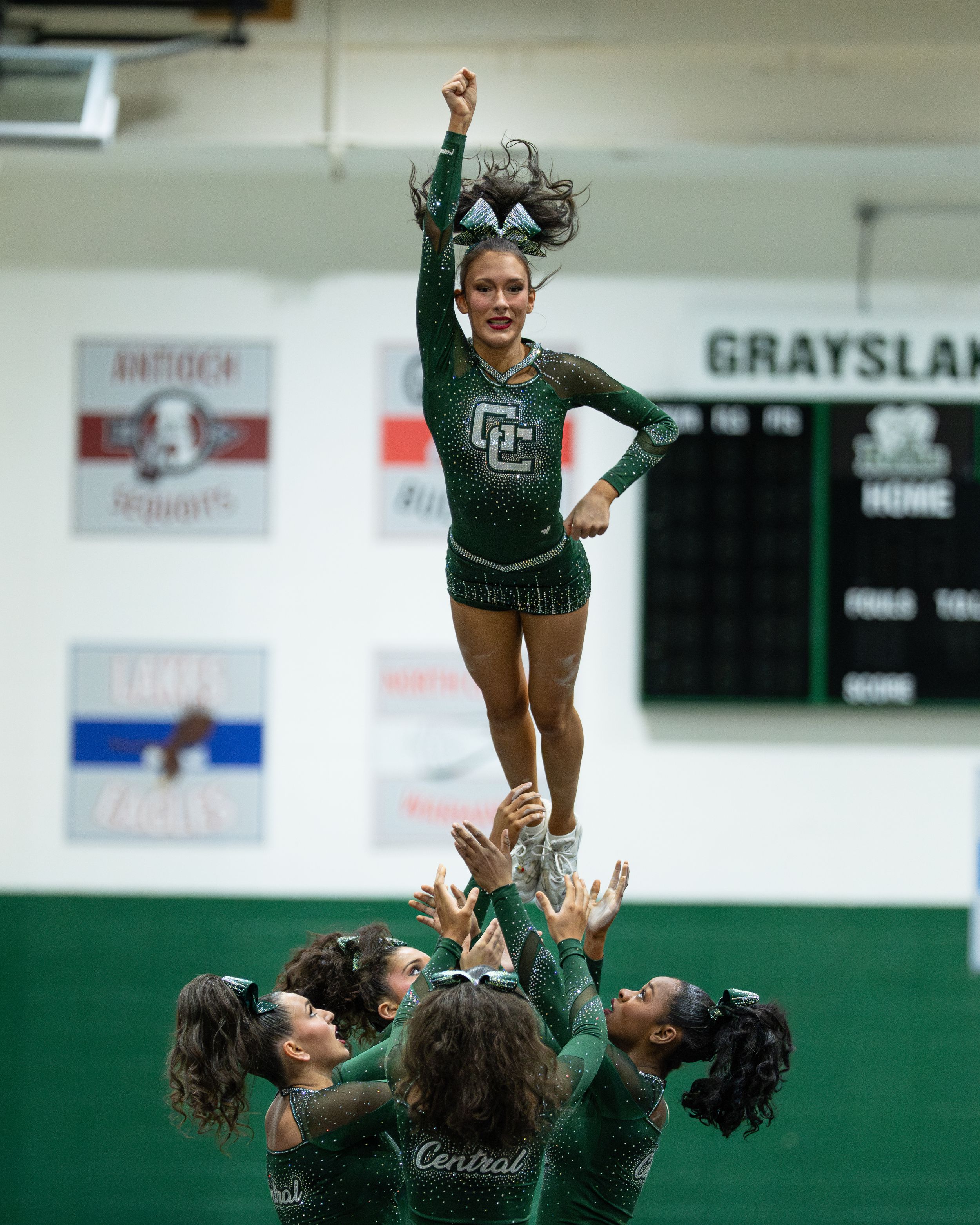 Cheerleader performs a high stunt lift at Grayslake Central competition, supported by teammates in green sparkle uniforms.