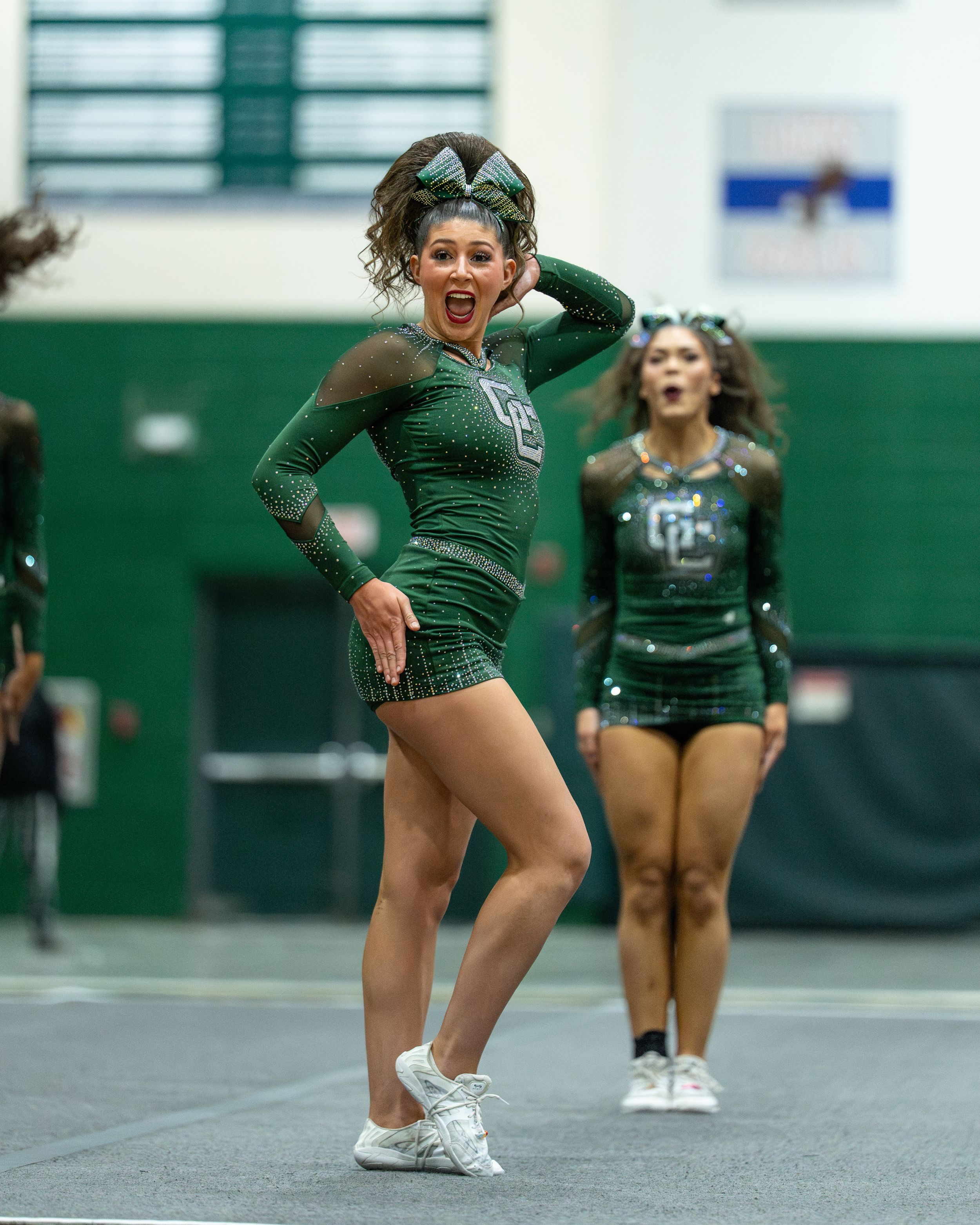 Two cheerleaders in green sequin uniforms perform an energetic routine during a competitive cheer event.