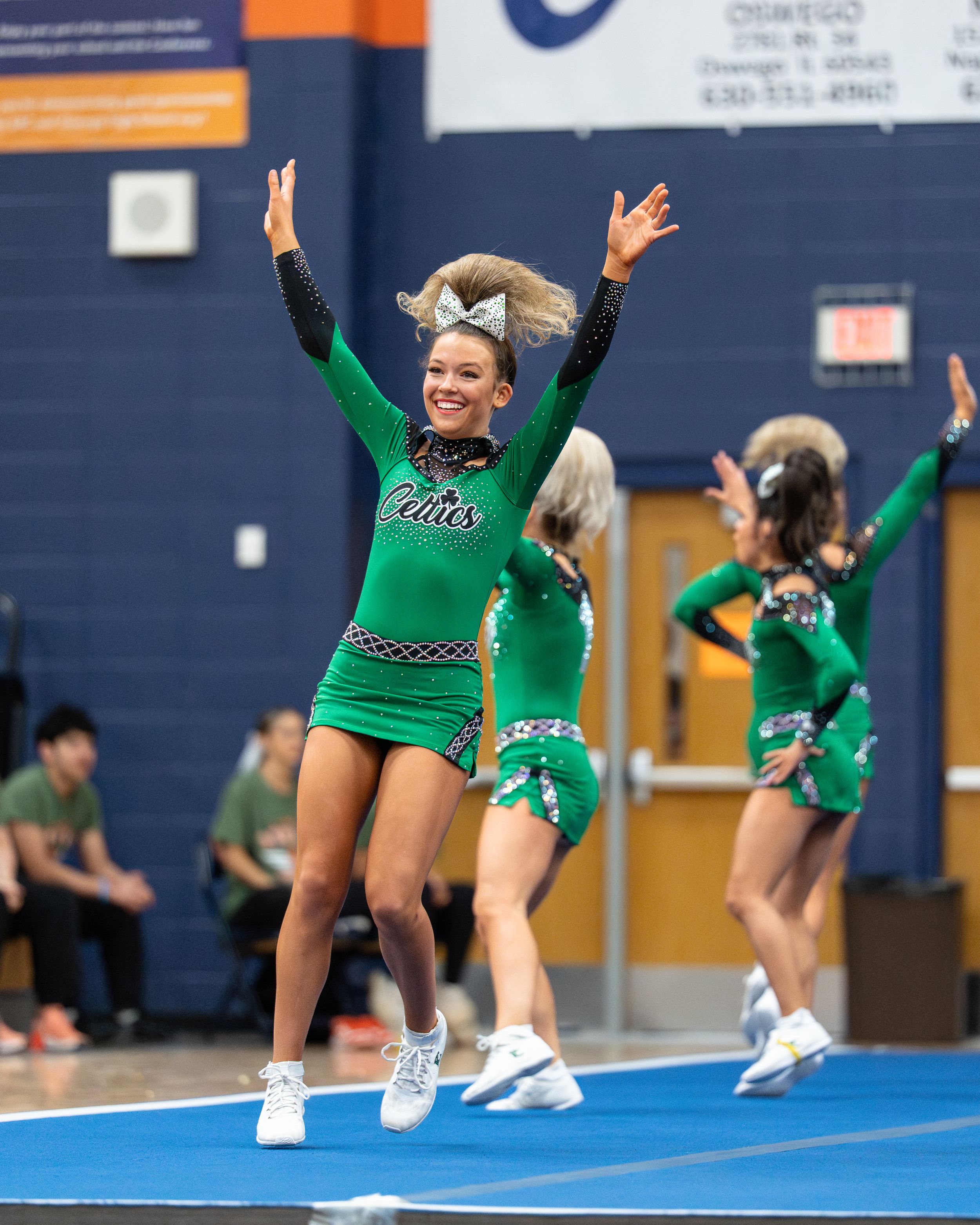 Smiling cheerleader in green uniform jumps with arms raised during a competitive cheerleading routine indoors.