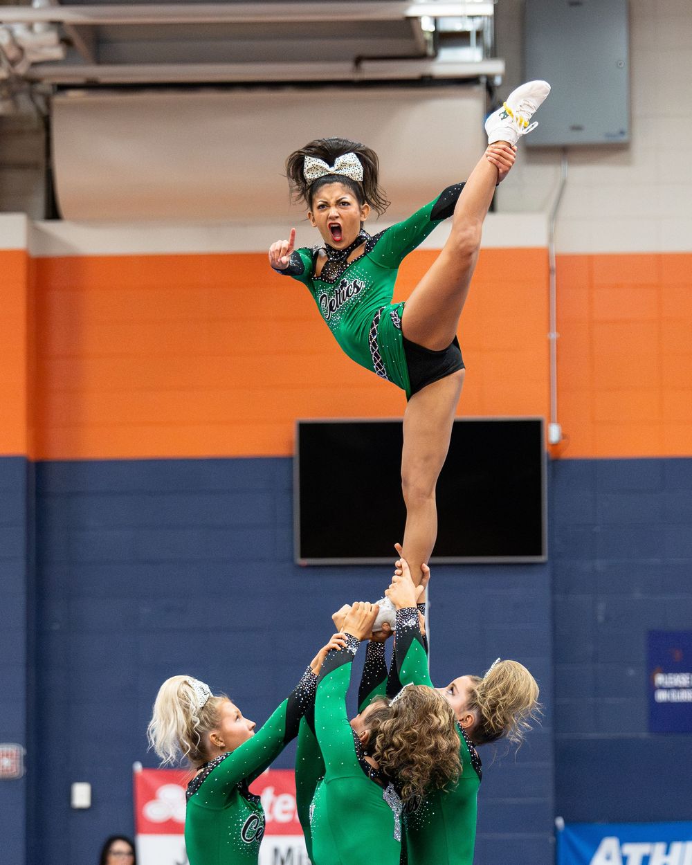 Cheerleaders in green uniforms perform a high-flying stunt, with one flyer executing a dramatic kick in a gymnasium.