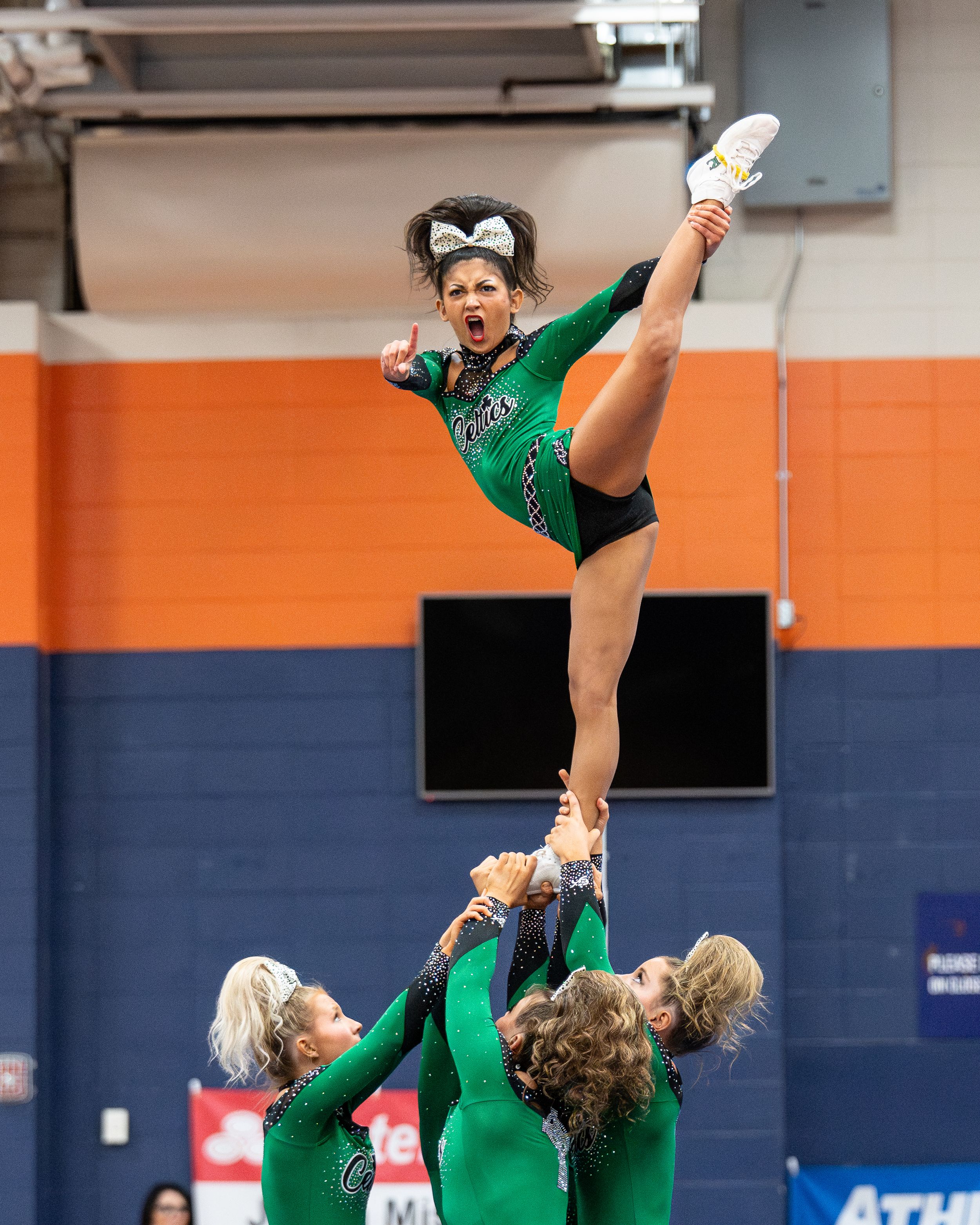 Cheerleaders in green uniforms perform a high-flying stunt, with one flyer executing a dramatic kick in a gymnasium.
