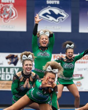 Cheerleaders in green Celtic uniforms perform energetically at a competition, with one flyer raising her arms in celebration.
