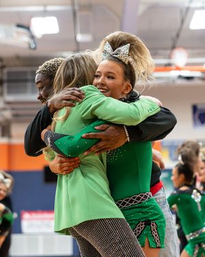 Cheerleaders in green uniforms share a warm group hug during a cheerleading competition inside a gymnasium.