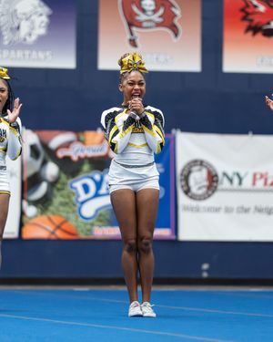 A cheerleader in black and gold uniform performs at a competition with Spartans banners in the background.