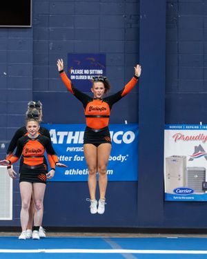 Two cheerleaders in orange and black uniforms perform at a competition, one jumping with arms raised.