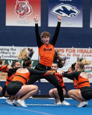 A male cheerleader in an orange uniform celebrates with arms raised while teammates kneel around him during a cheerleading competition.