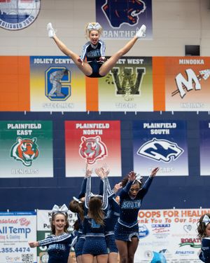 High school cheerleaders perform a stunt at a competition, with a flyer doing a toe-touch in the air above her teammates.