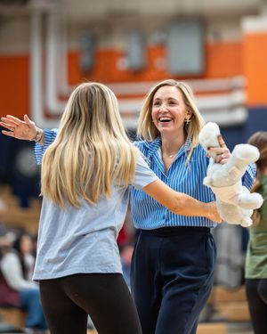 Two women in a gymnasium share an enthusiastic greeting, one holding balloon animals during a cheerleading event.