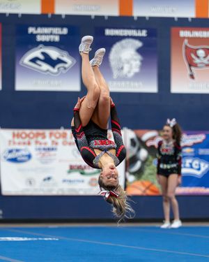 Cheerleader performing a backflip in mid-air at a high school cheerleading competition in a gymnasium.