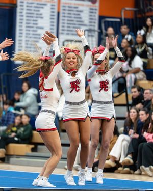 Three cheerleaders in white and red uniforms jump with arms raised during a competitive cheer performance.