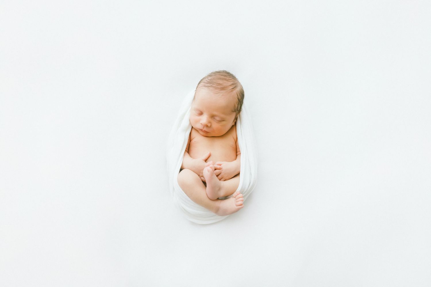 Peaceful newborn baby wrapped in white cloth, sleeping curled up on a white background in a professional photo.