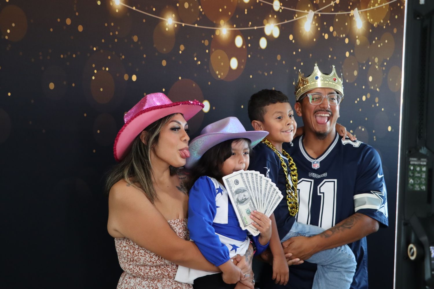Family of four posing at photo booth with gold bokeh backdrop, wearing cowboy hats and crown, holding fan of cash.
